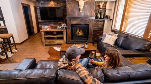 a couple relaxing on a dark leather couch drinking wine in front of a rustic fireplace with a skull mounted above it