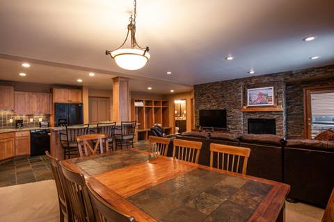 a spacious, open-concept large-scale lodge common area with a heavy wood dining table, stone accent wall, secondary fireplace, and kitchen bar stools