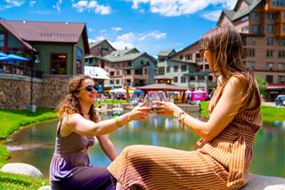 Two women sitting in front of a pond holding wine glasses