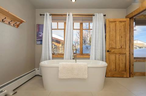Freestanding bathtub by large windows with mountain views, light curtains, and natural wood trim.