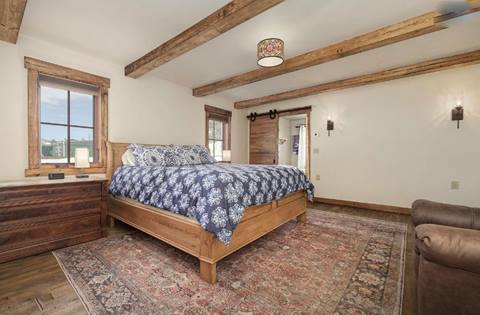 Master bedroom with king bed, exposed wood beams, large rug, and barn-style sliding door to en-suite bathroom.