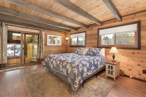 King bed with floral blanket and night stand next to it in room with wooded walls, windows, and a door to a patio. 