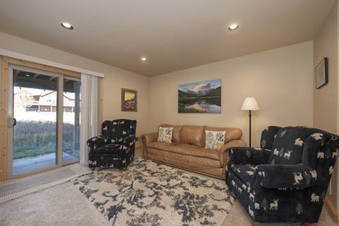 Cozy sitting area with a tan leather sofa, two navy armchairs with moose patterns, a patterned rug, sliding glass doors, and mountain-themed artwork.