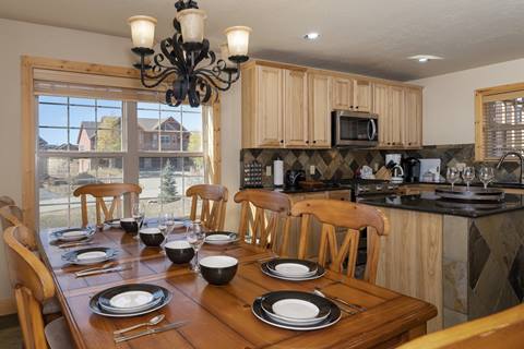 Dining area with large wooden table set for eight, next to kitchen with tile backsplash.