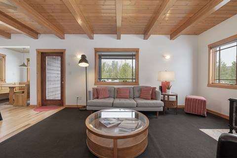 Living room with wood-beamed ceiling, gray sofa, striped pillows, glass coffee table, and bright window views.