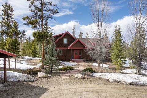 Exterior view of the rustic cabin with red trim, front porch, and snow-dusted yard surrounded by pine trees.