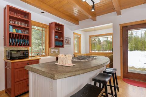 Rustic kitchen with red open cabinets, concrete island counter, and dining nook visible in background.