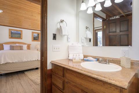 Bathroom vanity with wood cabinetry and countertop, sink, and mirror, looking into a queen bedroom.