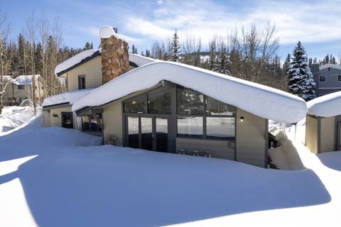 Snow-covered cabin exterior with large windows, stone chimney, and curved rooflines.