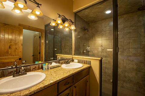 View of stone shower with warmly lit double sink. 