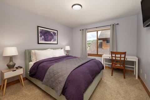 Bedroom with queen-size bed, purple bedding, nightstands, and a desk near the window.