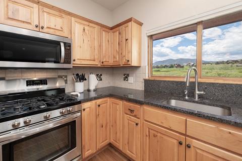 Stainless steel oven and microwave with wood cabinets, sink beneath a large window with scenic mountain views.
