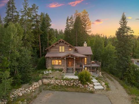 A rustic log cabin surrounded by pine trees under a colorful sunset sky, with warm light glowing from the windows.
