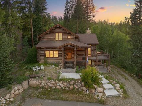 A log cabin nestled in a forest clearing during summer, with chairs on the lawn and golden evening light reflecting through the windows.