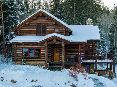 A wooden log cabin covered in snow, featuring a Colorado flag on the porch and trees blanketed in snow under a pink and orange winter sunset.