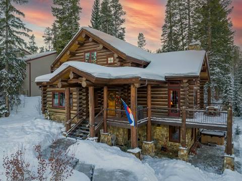 A front-facing view of a snow-covered log cabin with red-trimmed windows, surrounded by tall pine trees in a serene winter landscape.