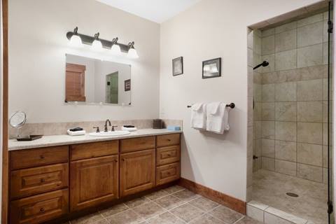 Spacious bathroom with large wooden vanity, double sinks, wall mirror with overhead lights, and tiled walk-in shower.
