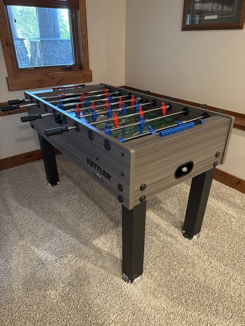 Kettler foosball table with red and blue players in a carpeted room with window and framed artwork.