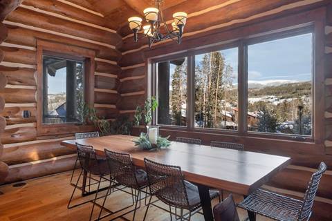 Large wooden dining table with eight chairs, log cabin walls, and wide windows overlooking snowy trees and mountains.