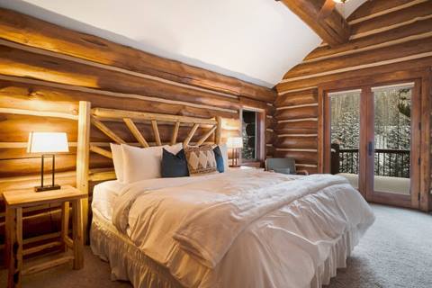 Log cabin bedroom with a king bed, rustic wooden headboard, nightstands, and glass doors opening to a snowy forest balcony.