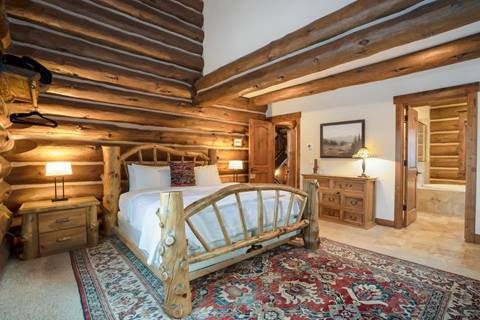 Log cabin bedroom featuring a natural wood bed frame, colorful patterned rug, dresser, and attached bathroom.