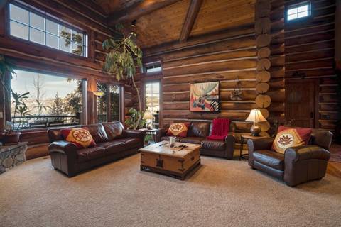 Large log cabin living room with leather sofas and armchairs, tall windows, rustic décor, and views of snowy trees.