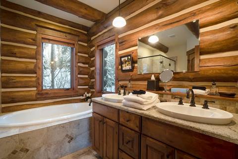 Bathroom with log walls, two sinks on a granite countertop, soaking tub by the window, and glass shower enclosure.