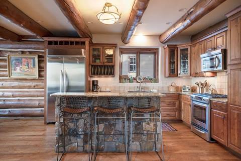 Rustic kitchen with wood cabinetry, granite island with stone base, three barstools, stainless steel appliances, and wood-beam ceiling.