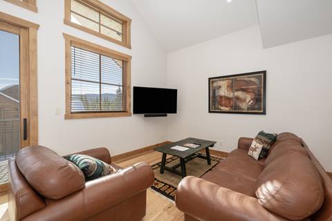 Cozy living area with leather sofas, mounted flat-screen TV, wall art, and natural light from large windows.