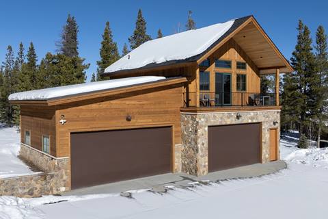 Exterior view of a large cabin with stone base, three-car garage, and snow-covered roofline.