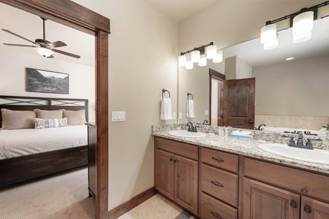 Double vanity bathroom with granite countertop and view into a bedroom with ceiling fan and framed landscape photo.