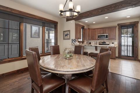 Dining area with round wooden table and leather chairs next to large windows.