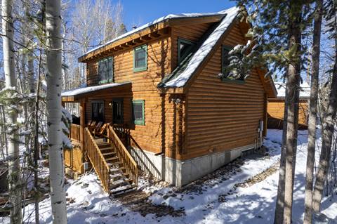 Exterior view of log cabin in winter with covered porch, stairs, and surrounding trees.