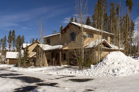 Two-story mountain home with stucco, timber accents, and snow-covered drive framed by tall pines.