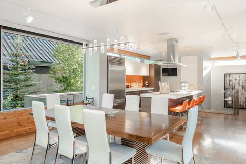 Dining area with modern wood table, white chairs, and view of kitchen with orange backsplash.