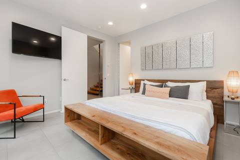 Modern bedroom with wood-frame king bed, white bedding, orange accent chair, wall-mounted TV, and contemporary artwork.