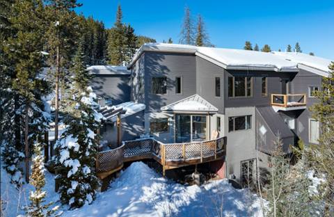 Winter exterior of the same mountain home, covered in snow, with a wraparound deck and forest backdrop.