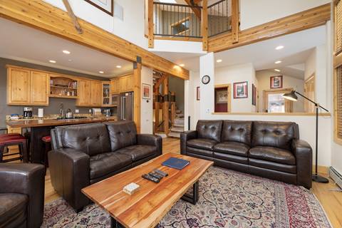 Living room with leather seating opening to a kitchen with wooden cabinetry and center island, staircase in the background.