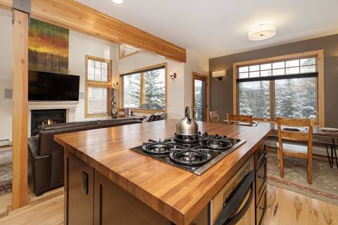 Kitchen island with gas cooktop and kettle, open view into living and dining rooms with forest views outside.